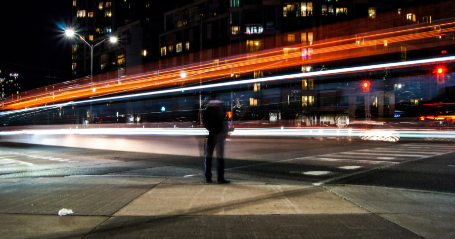 man standing next to city street at night with red and white lights streaming by using a long-exposure photo effect