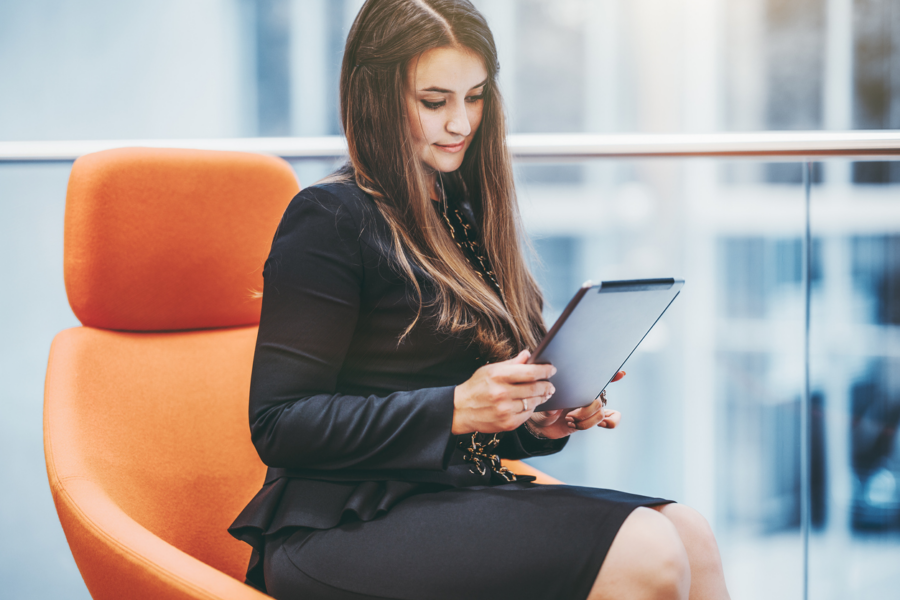 Woman with brown hair and black skirt and top sitting on a plush orange chair looking contently at a tablet in her hands