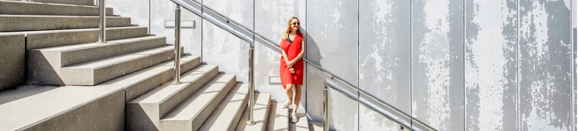 A woman in a red dress and glasses standing on stairs in front of a gray wall.