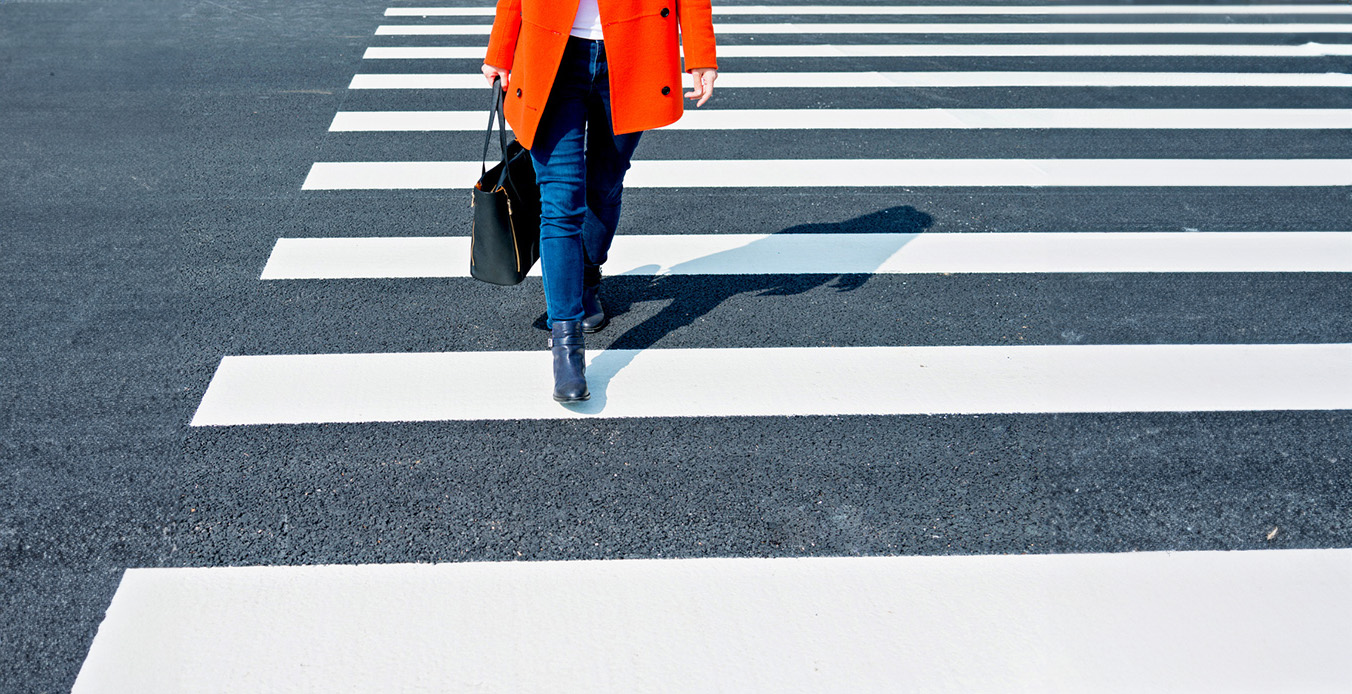 Woman in jeans and orange jacket crossing a road.