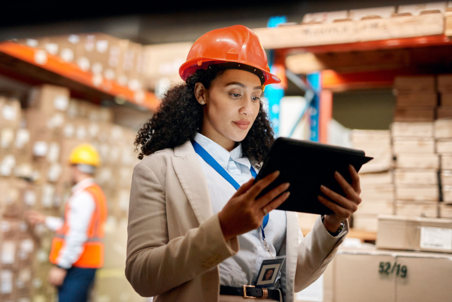 African American woman wearing orange hardhat looking thoughtfully at a tablet in a large warehouse distribution center